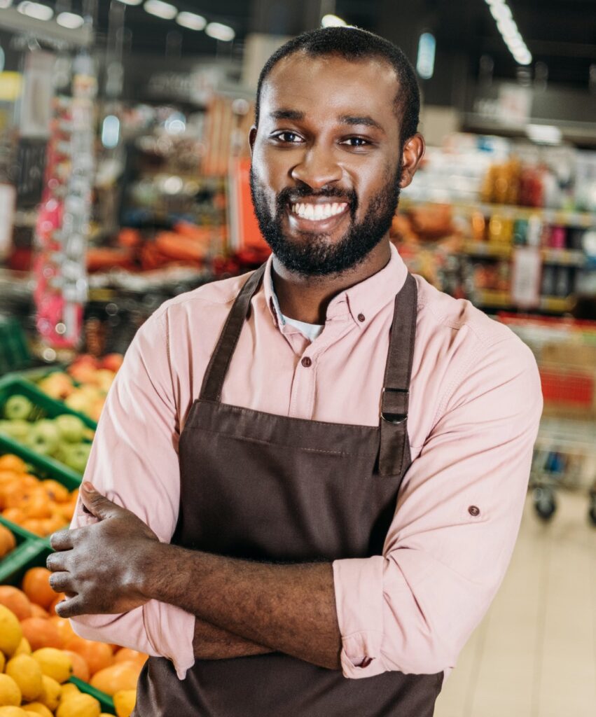 african american male shop assistant in apron standing with crossed arms near fruit department in